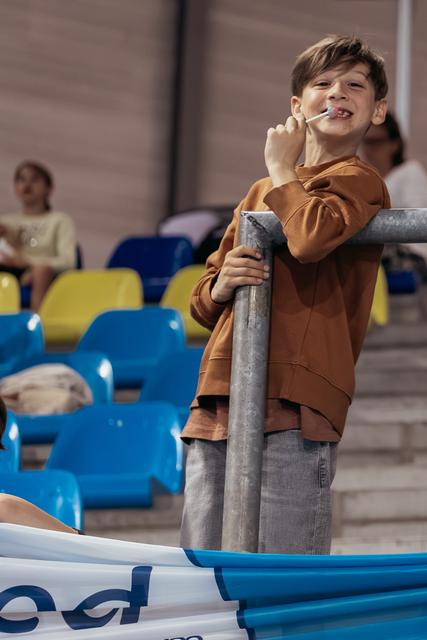 Young boy grins while eating a lollipop, leaning on a metal railing in colorful stadium bleachers.