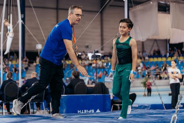 Coach in blue leans toward young gymnast in green uniform, sharing a warm interaction on the trampoline floor at indoor arena.