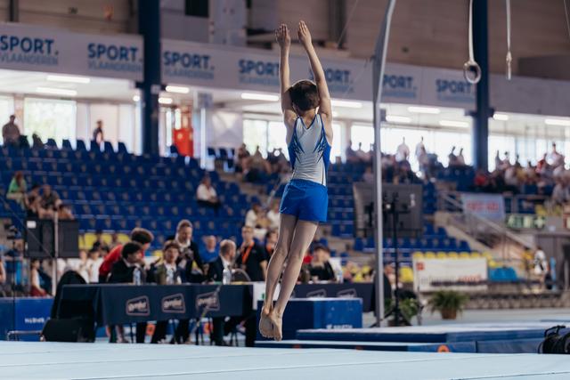 Young gymnast mid-air with arms raised overhead, landing on floor exercise mat in a packed indoor arena.