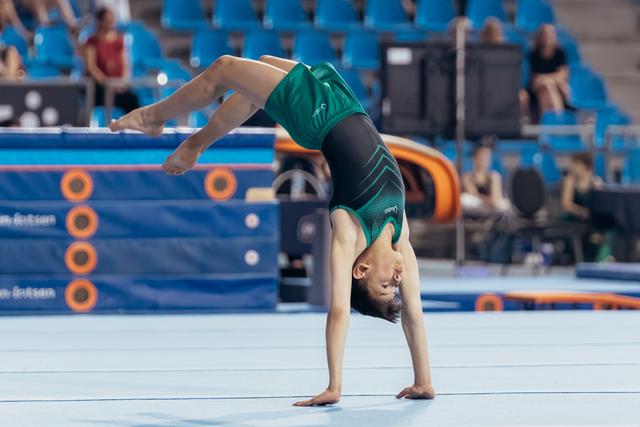 Young gymnast performs a backbend on the floor exercise mat during a competition, legs extended mid-movement.