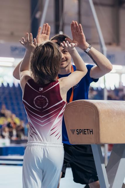 Young gymnast and smiling coach share an enthusiastic high-five beside a Spieth pommel horse in a gymnastics hall.