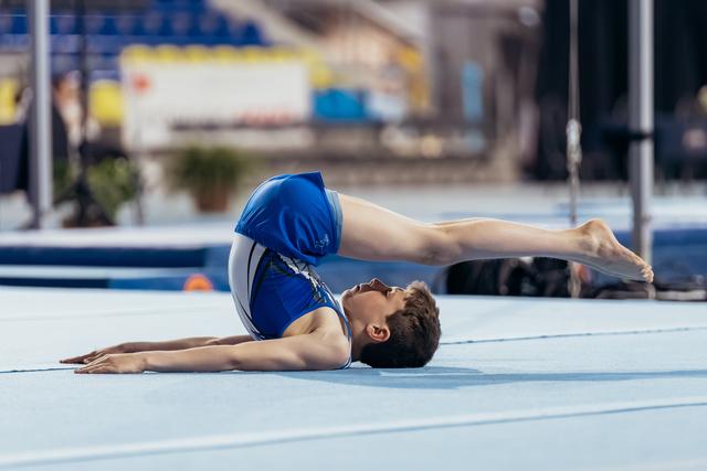 Young male gymnast performs a bridge pose on floor exercise mat, upper body stretched flat, legs extended upward.