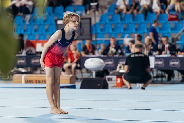 Young gymnast stands barefoot on floor mat, leaning forward with intense focus, preparing for her routine at a gymnastics competition.