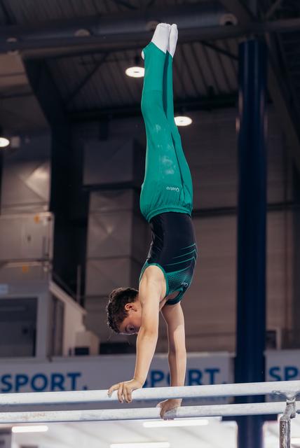 Young male gymnast performs a precise handstand on parallel bars, wearing teal and black attire, in an indoor gymnasium.