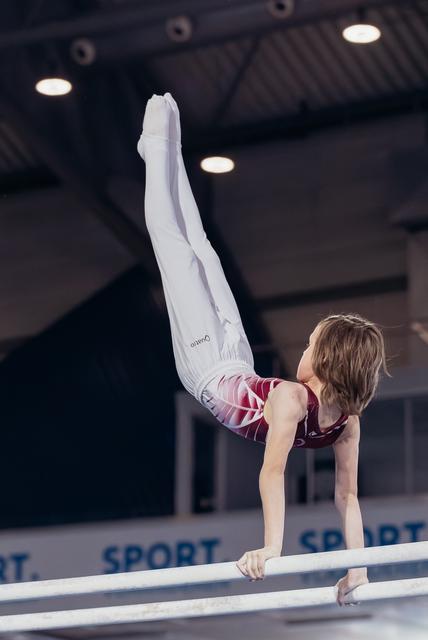 Young gymnast performs a precise handstand on parallel bars, legs extended upward, in a lit sports hall.