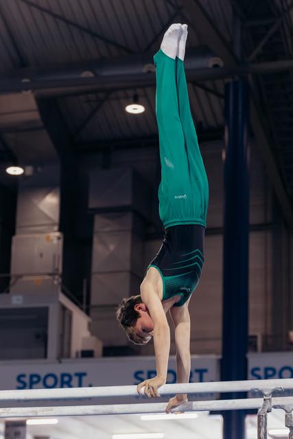 Young male gymnast holds a perfect handstand on parallel bars, legs straight up, wearing teal outfit in indoor gym.