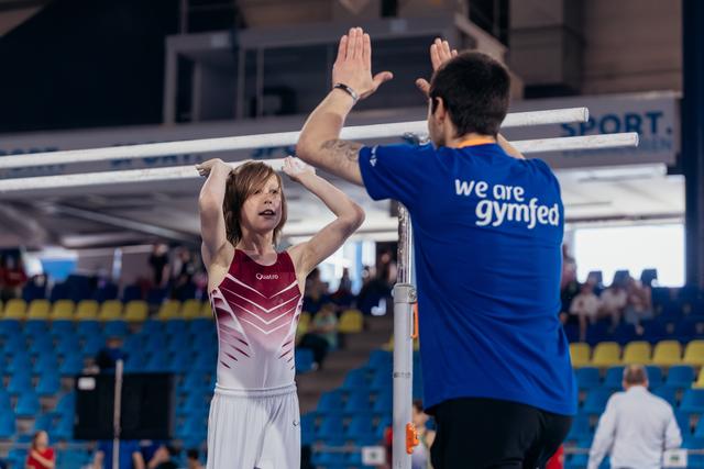 Young gymnast in red leotard high-fives her coach after uneven bars routine, both arms raised in celebration.