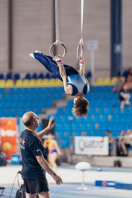 Young gymnast hangs inverted on still rings while attentive coach below reaches up to guide and spot during an indoor gymnastics event.