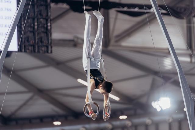 Young male gymnast holds an inverted cross position on still rings, face showing intense concentration in a competition hall.