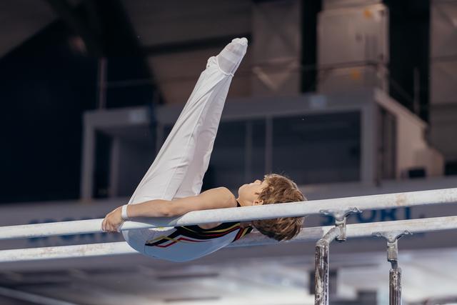 Young male gymnast performs a back arch on parallel bars, legs extended upward, showcasing strength and flexibility.