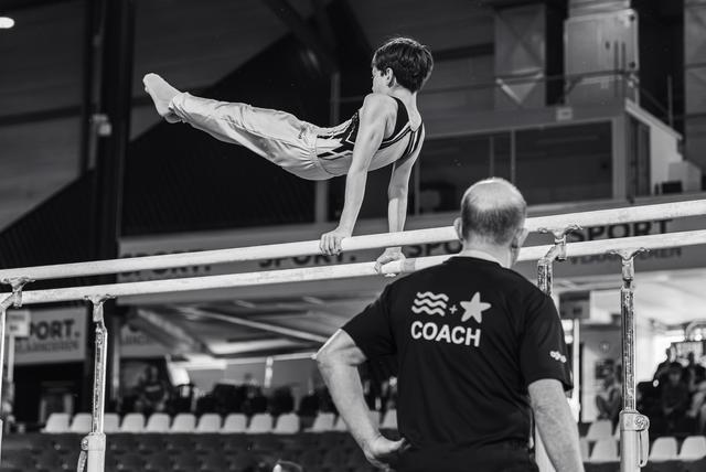 Young male gymnast performs a split hold on parallel bars while his coach observes attentively from below at a competition.