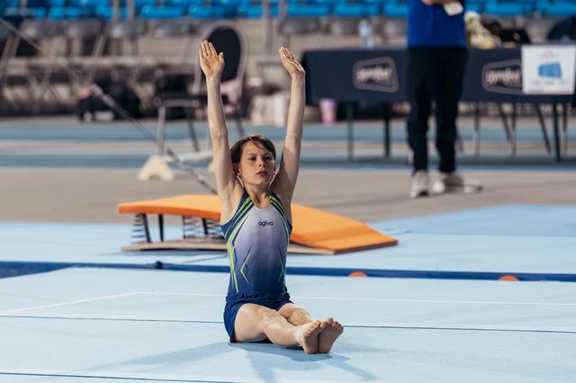 Young gymnast sits on blue floor mat, arms raised high with focused expression, wearing blue agiva leotard.
