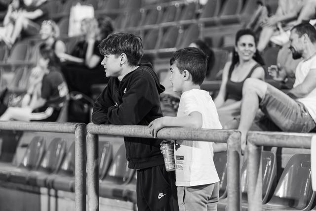 Two boys lean on a railing at a stadium, chatting while watching a sports event, one holding a drink can.