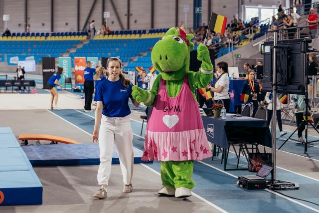 Smiling woman in blue shirt walks hand-in-hand with green GYM mascot waving Belgian flag at indoor gymnastics event.