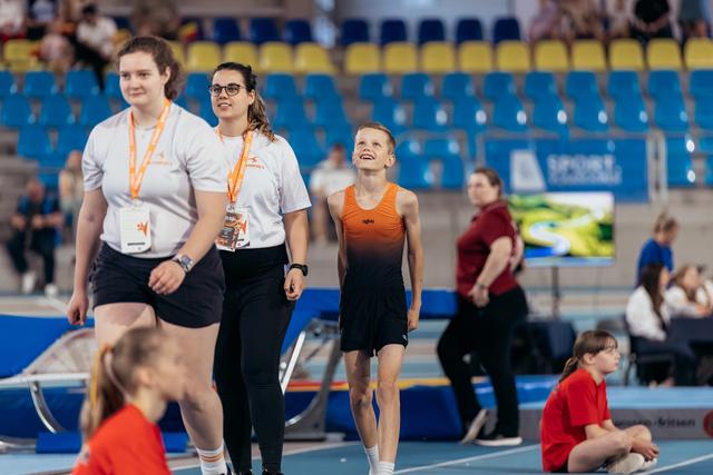 A smiling young gymnast in orange walks alongside two coaches in white shirts with lanyards on an indoor track.