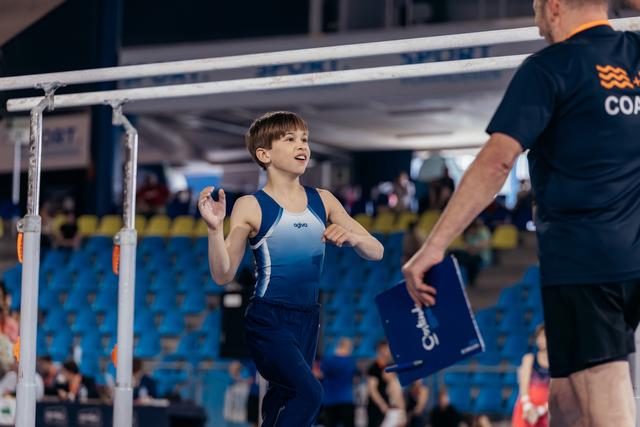 Young male gymnast walks toward coach with an eager expression, parallel bars behind him in a competition arena.