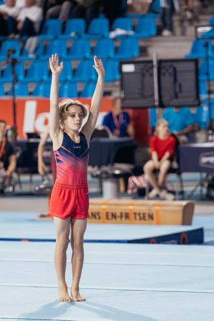 Young gymnast stands barefoot on the mat, arms raised high in a confident salute, wearing a red and navy leotard.