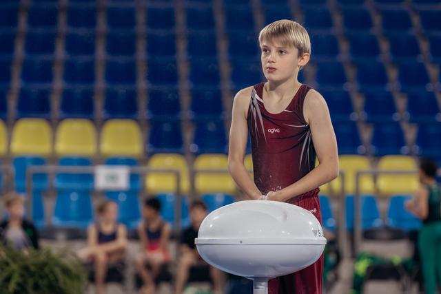 Blonde boy gymnast in dark red leotard stands at chalk bowl, gazing intently aside in a competition arena.