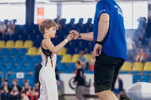 Young gymnast in white uniform fist-bumps coach in blue shirt, sharing a moment of encouragement in a busy sports arena.