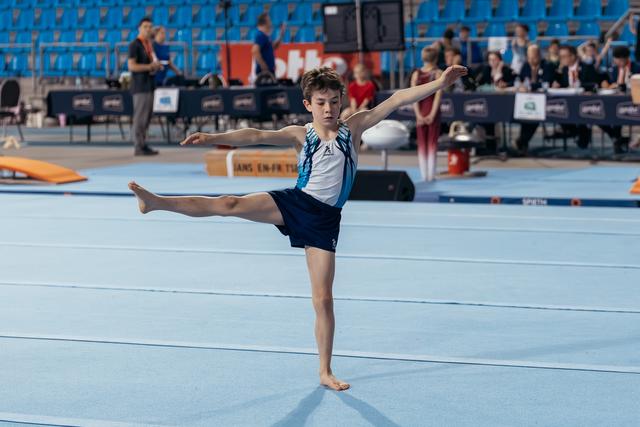 Young male gymnast performs an arabesque on the floor exercise, arms outstretched, focused expression in a competition arena.