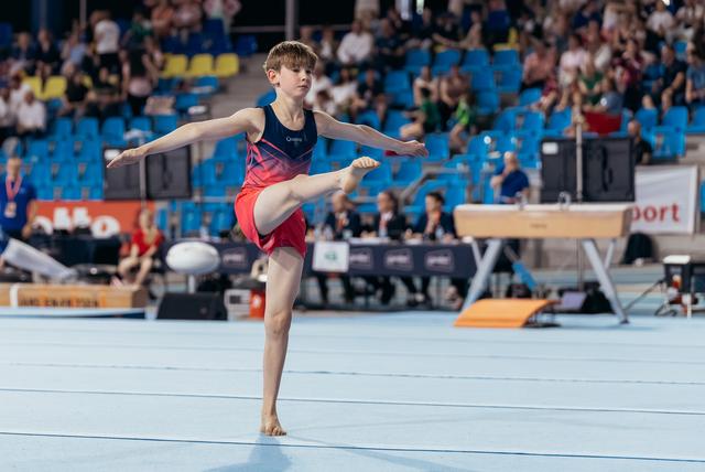 Young male gymnast balances on one leg with arms outstretched during a floor exercise routine at an indoor arena.