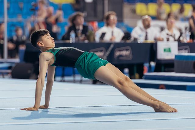 Young male gymnast performs a graceful floor routine, holding a split-leg pose with focused expression at a gymnastics meet.