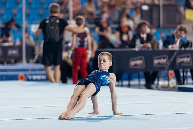 Young boy gymnast performs a floor routine, holding a low arabesque position with intense focus on his face.