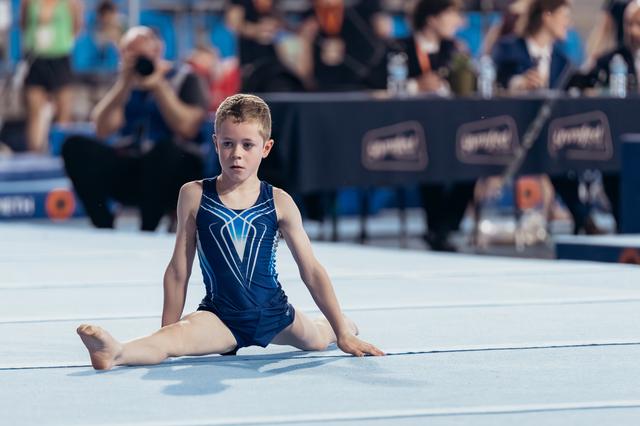 Young boy gymnast sits in straddle position on floor mat, expression focused and composed during competition.