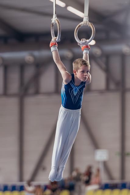 Focused young male gymnast hangs from still rings, legs together, intense concentration, in a gymnasium during competition.