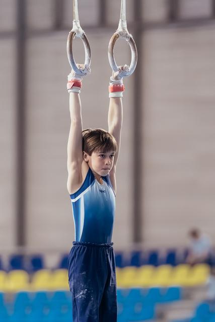 Focused young male gymnast hangs from still rings, arms raised overhead, wearing blue leo in a sports hall.
