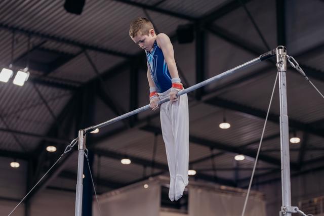Young male gymnast grips the high bar with intense focus, body suspended mid-routine in an indoor gymnasium.