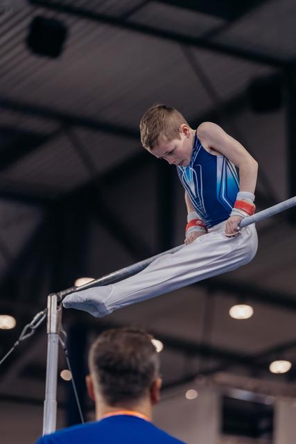 Young boy gymnast grips the high bar with intense focus, legs extended, wearing blue leotard and wrist guards indoors.