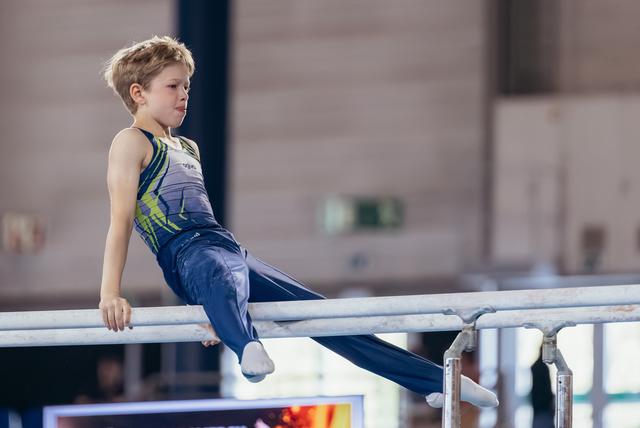 Focused young male gymnast seated on parallel bars, legs extended, wearing blue and green leotard in indoor gymnastics hall.