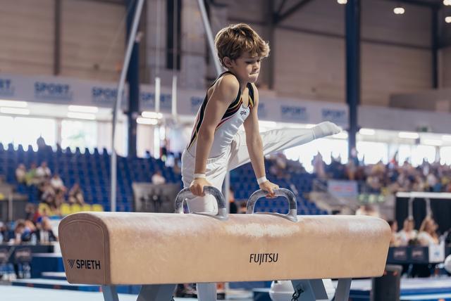 Young male gymnast stands concentrated on a Spieth pommel horse during an indoor gymnastics event, gripping the handles.