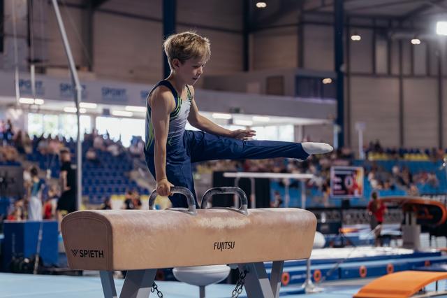 Focused young male gymnast performs on pommel horse, leg extended, in a busy competition arena.
