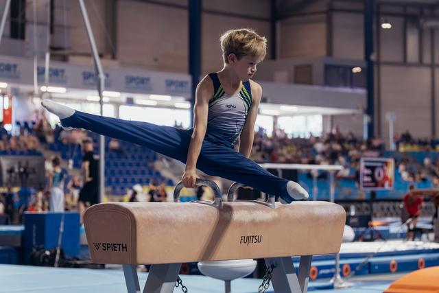 Focused young male gymnast executes a scissor split on a Spieth pommel horse during a competitive gymnastics event.
