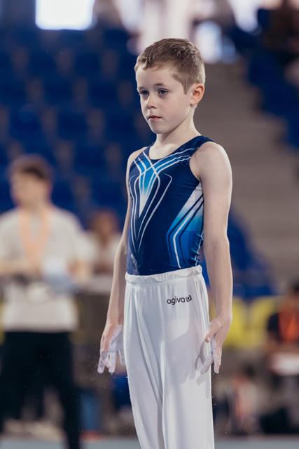 Young male gymnast stands composed and focused, wearing a navy blue leotard and white pants, chalked hands at sides, in a gymnasium.