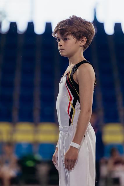 Young male gymnast stands still, focused expression, wearing white and black Belgian uniform in a competition arena.