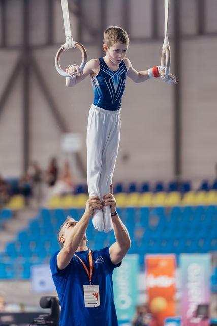 Focused young gymnast holds still rings while coach steadies his feet in a blue-seated sports arena.