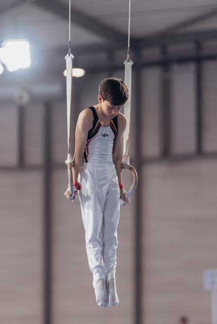 Young male gymnast hangs still on rings, head bowed in concentration, wearing white and black competition leotard indoors.