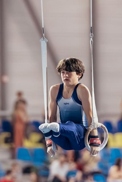Young male gymnast performs an L-sit on still rings, expression focused and determined, in a competition gymnasium.