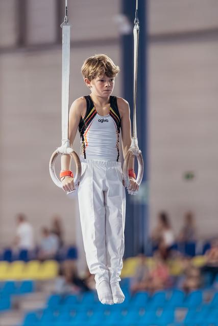 Young male gymnast holds still rings with focused concentration, wearing white and black agiva leotard in indoor arena.