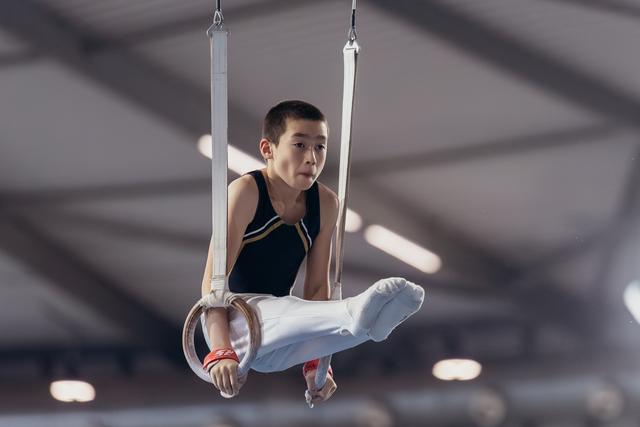 Young male gymnast holds a tucked position on still rings, expression focused and determined, inside a gymnasium.