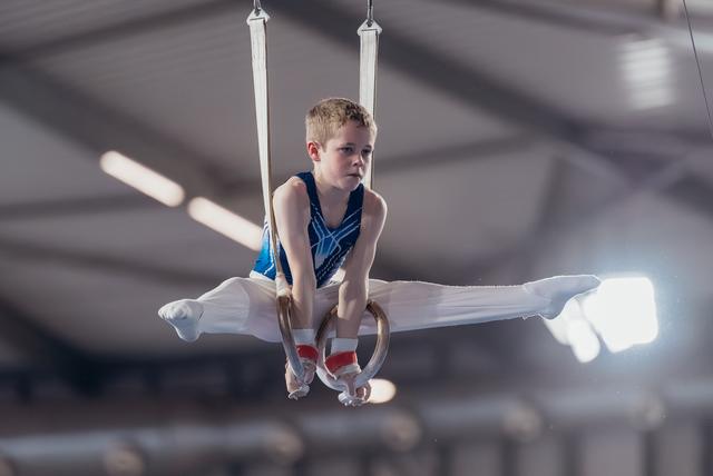 Young male gymnast holds a tuck position on still rings, wearing a blue leotard, with intense concentration.
