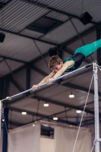 Young male gymnast grips the horizontal bar with fierce concentration, body stretched horizontal in a gymnastics hall.