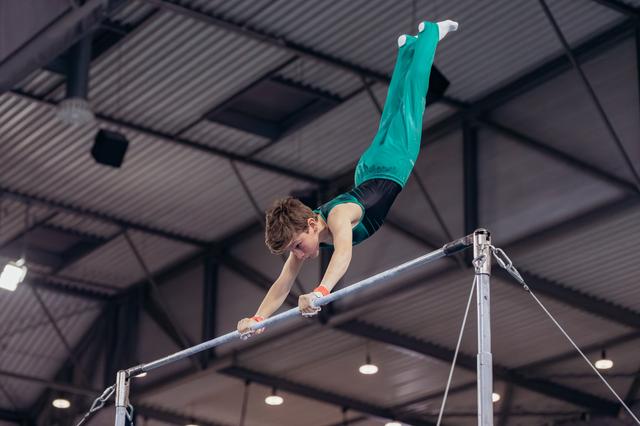 Young male gymnast grips the high bar with fierce concentration, body extended horizontally in a dynamic gymnastics hall.