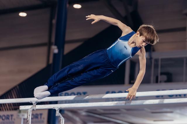 Young male gymnast performs a powerful hold on parallel bars, body extended horizontally, focused expression in indoor gym.