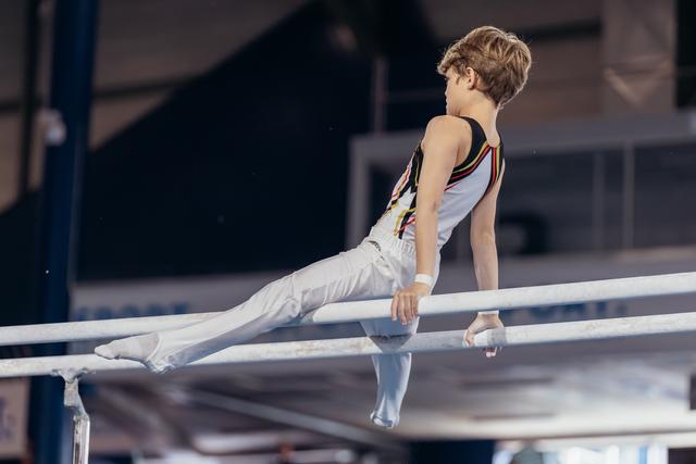 Young male gymnast holds a straddle position on parallel bars, showing concentration and strength in a gym.
