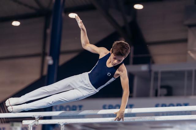 Young male gymnast performs a precise horizontal hold on parallel bars, one arm raised, showing intense focus.