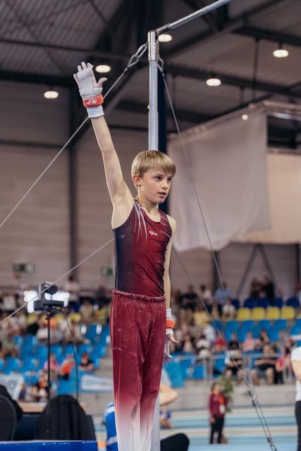 Young male gymnast raises hand confidently before high bar routine in a packed indoor gymnastics arena.